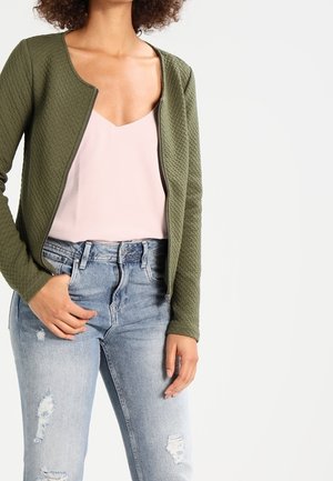 Vrouw draagt een olijfgroene gestructureerde jas met ritssluiting, lichtroze hemdje en lichtblauwe gescheurde spijkerbroek, staand met één hand in de zak.