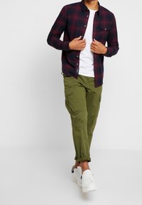 Camicia a quadri blu navy e bordeaux, abbinata a pantaloni cargo verdi oliva. Maglietta bianca sotto e sneakers bianche completano il look.