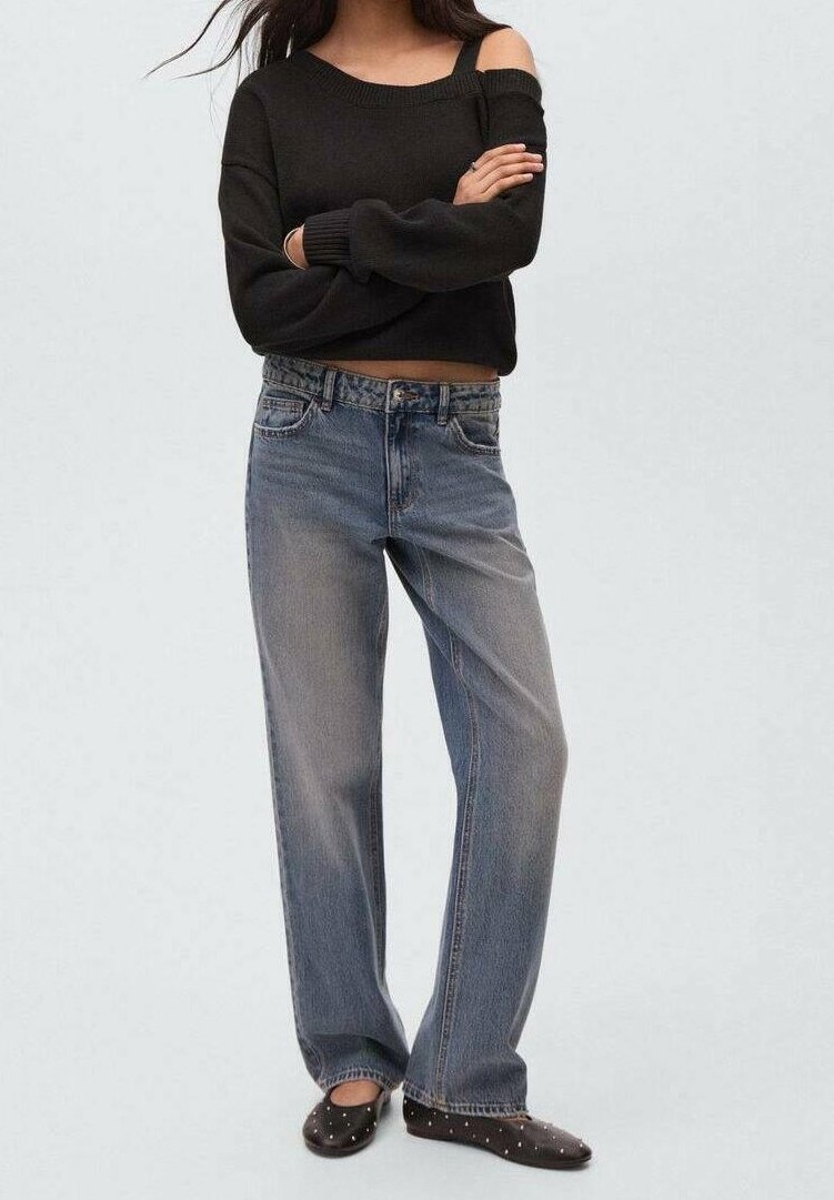Femme debout, les bras croisés, portant un pull noir épaules dénudées, un jean bleu délavé et des ballerines noires cloutées.