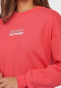 Sweatshirt in cotone corallo con colletto e polsini a costine. Presenta la scritta bianca "The Future Surf Society" in un design centrato.