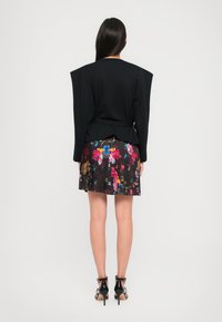 Zwarte oversized top met brede schouders, gecombineerd met een geplooide rok met een bloemenpatroon in levendige kleuren. De hakken zijn glanzend zwart.