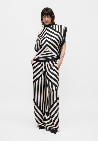 Γυναίκα που φοράει φαρδιά παντελόνια με ασπρόμαυρες γεωμετρικές ρίγες και αντίστοιχο φαρδύ τοπ, στέκεται με τα χέρια στις τσέπες μπροστά σε ένα απλό φόντο.