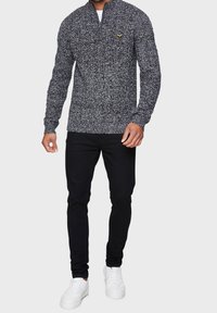 Gestrickter Kapuzenpullover in Schwarz und Weiß mit einer leichten Textur und einem kleinen gelben Logo. Kombiniert mit engen schwarzen Hosen und weißen Sneakers.