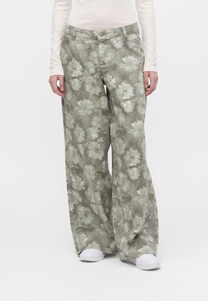 Personne portant un pantalon vert à jambes larges avec imprimé floral blanc, un haut blanc à manches longues et des chaussures blanches, debout devant un fond uni.