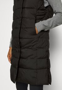 Personne portant une longue veste matelassée noire avec les mains dans les poches latérales, par-dessus un t-shirt gris à manches longues, sur un fond uni.