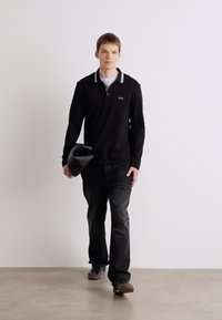 Camisa polo preta de manga longa com colarinho branco, combinada com jeans escuros e sapatos. A figura segura um chapéu preto com uma das mãos.