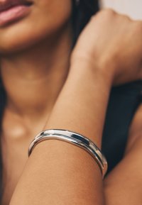 Silberfarbenes Metall-Armband liegt auf einem braunhäutigen Unterarm, mit einer glatten, glänzenden Oberfläche und schlichtem, abgerundetem Design.