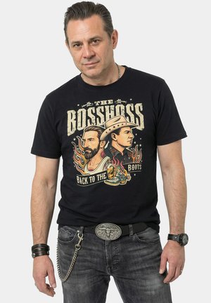 Man draagt een zwart T-shirt "The Bosshoss Back to the Boots" met geïllustreerde profielen, gecombineerd met vervaagde jeans en accessoires.