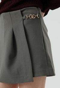 Jupe plissée grise avec une texture lisse, agrémentée d'un détail en chaîne dorée à la taille. Le tissu est léger et tombe élégamment.