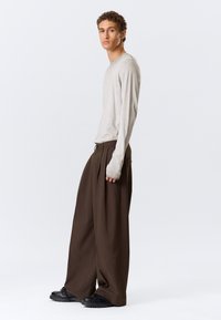 Chemise beige à manches longues associée à un pantalon large marron et à des baskets noires épaisses. Coupe décontractée avec coutures visibles et silhouette ample.