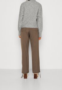 Grau gestrickter Pullover mit hohem Kragen, kombiniert mit braunen weit geschnittenen Hosen. Die Hose hat Taschen auf der Rückseite und gebundene Säume. Braune Blockabsatzschuhe.