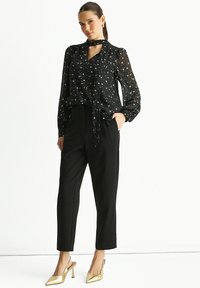 Black star-patterned blouse with a tie neck, sheer fabric, and long sleeves, paired with high-waisted black trousers and gold heels.