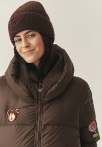 Barna pufi dzseki nagy kapucnival, amelyen egy medve folt és egy hímzett virág található. Finoman bordázott barna kötött gallér alatta.