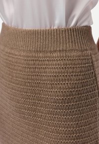 Gros plan sur une personne portant une jupe marron en maille côtelée et texturée, associée à un chemisier en satin blanc rentré.