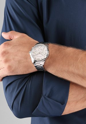 Montre chronographe argentée avec un cadran blanc texturé, un bracelet en métal noir et argent, et trois sous-cadrans, portée sur un bras bronzé.