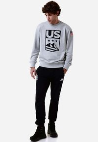 Felpa grigio chiaro con un grande logo nero "US" con montagne e stelle. Abbinata a pantaloni jogger blu navy e calzature nere.