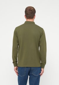 Oliwkowa, długorękawowa koszulka polo z kołnierzykiem, gładka w dotyku i z prążkowanymi mankietami, noszona z niebieskimi dżinsami, widok z tyłu.