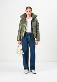 Vrouw in een olijfgroene pufferjack, witte shirt, blauwe jeans, witte schoenen, die een kleine beige handtas vasthoudt tegen een plain witte achtergrond.