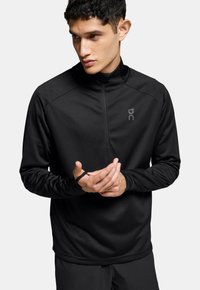 Haut de sport noir à manches longues avec col montant, fermeture éclair mi-hauteur à l'avant, ouvertures pour les pouces et tissu texturé avec un subtil accent logo.