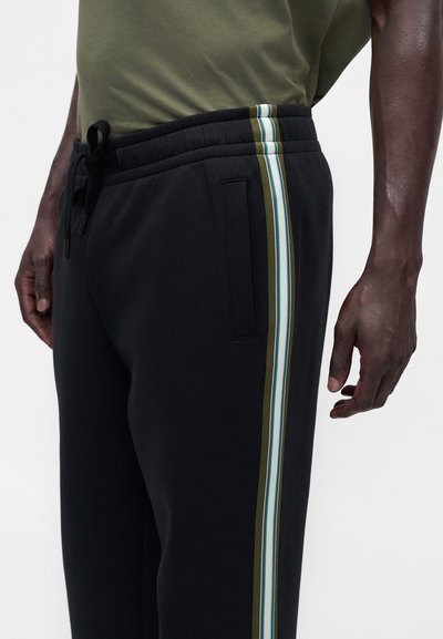 Homme portant un pantalon noir avec des bandes latérales verticales olive, blanche et bleue et une taille à cordon, associé à une chemise vert olive.