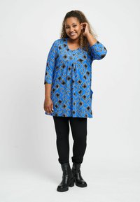 Niebieska tunika w wzory z czarnymi akcentami, z delikatnym zakładkiem i rękawami o długości trzech ćwiartek, w połączeniu z czarnymi legginsami i botkami za kostkę.