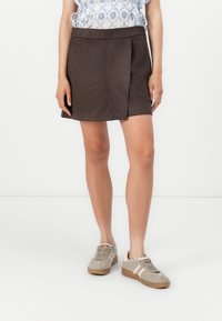 Mini gonna in suede marrone con spacco frontale, abbinata a sneakers chiare e una blusa blu con motivo floreale.