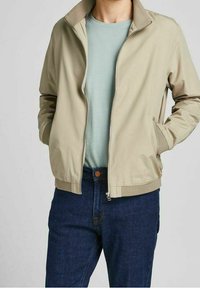 Světlý béžový bomber s zipem, bočními kapsami a kontrastním texturovaným límcem. Kombinován s světle modrým tričkem a tmavými džínami.