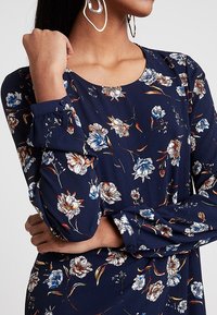 Abito floreale blu navy con maniche lunghe, caratterizzato da fiori bianchi, blu e arancioni. Tessuto morbido e scollatura rotonda con polsini con bottoni.