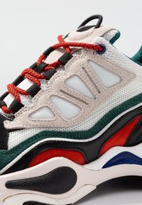 Flerfärgade sneakers i nät och mocka, med röda och svarta snören. Märkbara detaljer inkluderar texturerade överlägg och en tjock sula.