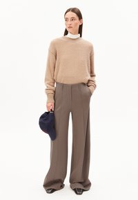 Beige gestrickter Pullover mit rundem Ausschnitt, kombiniert mit hochtaillierten, weit geschnittenen braunen Hosen. Hält eine dunkelblaue Mütze.