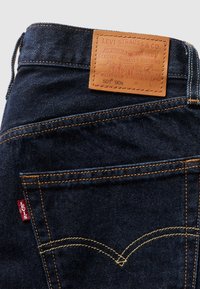 Poche arrière de jean Levi’s bleu foncé avec surpiqûre jaune, écusson de marque en cuir fauve et étiquette Levi’s rouge sur fond clair.