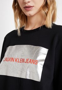Felpa nera con una fascia rettangolare argentata testurizzata, caratterizzata da pieghe, sulla quale è stampato "CALVIN KLEIN JEANS" in lettering arancione vivace.