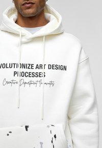 Witte hoodie met een trekkoord, voorzien van zwarte tekst en een verfspetters ontwerp in grijs en blauw op de onderkant van de voorkant. Zachte stofstructuur.