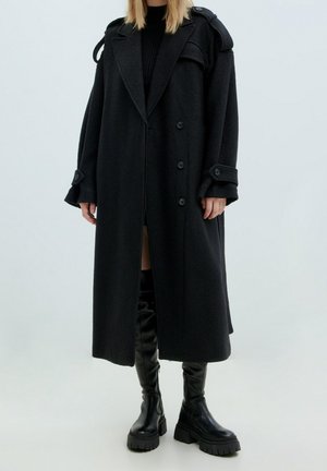Lange zwarte wollen jas met een dubbelknoopsluiting, grote knopen en schouderaccenten, gecombineerd met zwarte laarzen tot de knie.