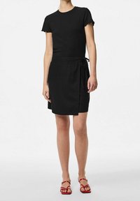 Černé žebrované tričko s krátkými volánkovými rukávy v kombinaci s černou zavinovací sukní se zavázaným pasem, doplněné červenými sandály na šněrování.