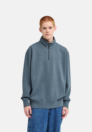 Jonge persoon met kort rood haar die een losse blauwe trui met een kwartritssluiting en blauwe jeans draagt, staand tegen een effen achtergrond.