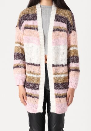 Frau trägt einen flauschigen, mehrfarbig gestreiften Cardigan in Rosa-, Weiß-, Braun- und Beigetönen über einem grauen Crop-Top und schwarzen Hosen.