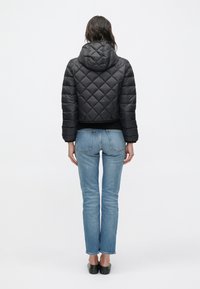 Άτομο που φοράει μαύρο καπιτονέ μπουφάν με κουκούλα, μπλε τζιν και μαύρα παπούτσια slip-on, στέκεται με την πλάτη γυρισμένη σε απλό λευκό φόντο.