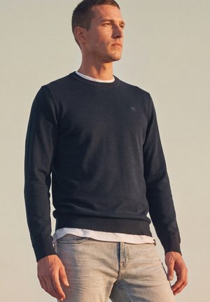 Junger Mann steht im Freien, trägt einen dunklen Langarmpullover über einem weißen Hemd und hellblaue Jeans und schaut zur Seite.