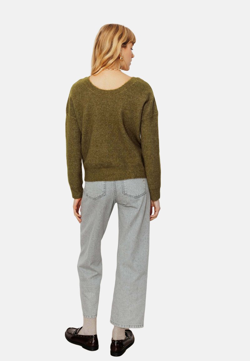 Pull en maille vert olive avec un large col, associé à un jean gris clair à jambes droites. Le mannequin porte des chaussures sombres et des chaussettes.