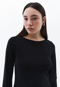Schwarzes Langarmoberteil mit rundem Ausschnitt, das dezente Nähte und eine glatte Textur aufweist. Das Model trägt goldene Creolen.