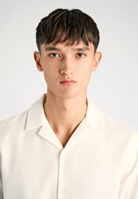 Camisa de botón de manga corta, color beige claro, con cuello, tela texturizada y un sutil detalle de logo en la parte superior izquierda. Diseño suave y limpio.