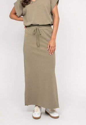 Femme portant une jupe maxi en maille beige avec taille à cordon et des baskets blanches, debout devant un fond blanc uni.