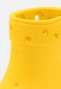 Stivaletto di gomma giallo con superficie testurizzata, fori rotondi per ventilazione e bordo superiore arrotondato con logo impresso.