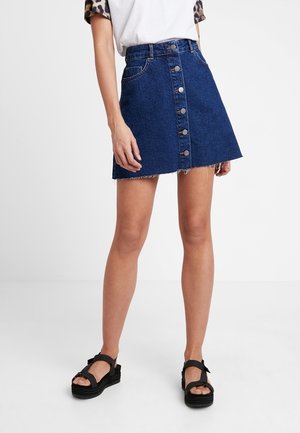 Femme portant une mini-jupe en jean bleu foncé à boutons sur le devant et des sandales compensées noires, debout sur un fond blanc uni.