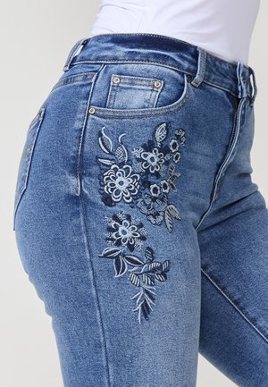 Jean en denim bleu clair avec un motif floral brodé sur le côté gauche. Il dispose de cinq poches et d'une silhouette ajustée.