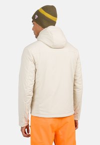 Doudoune beige clair avec capuche, présentant des coutures surpiquées et une texture lisse, associée à un bonnet marron avec des rayures jaunes.