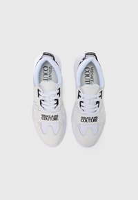 Białe trampki wykonane z połączenia gładkiej skóry i siateczki. Charakteryzują się wyraźnym logo na pasku, płaskimi sznurowadłami i grubą gumową podeszwą.