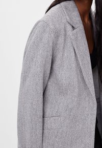 Grauer Blazer mit Hahnentrittmuster, strukturiertem Design, Reverskragen und leicht strukturierter Stoffstruktur. Verfügt über eine Tasche an der Vorderseite.