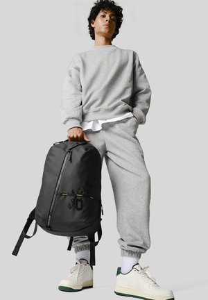 Felpa grigia e pantaloni felpati con polsini elasticizzati, abbinati a uno zaino nero con dettagli gialli e cintura frontale con zip. Sneakers bianche.